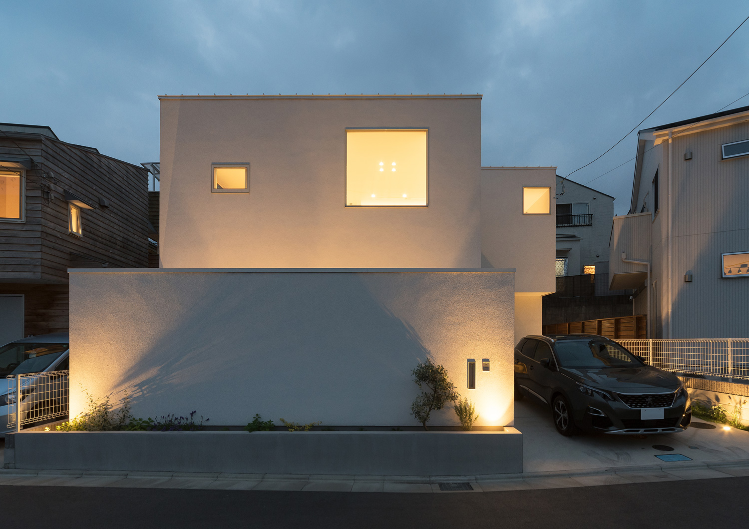 白い外壁が照明に照らされ正方形の窓から光が漏れる住宅・デザイン住宅