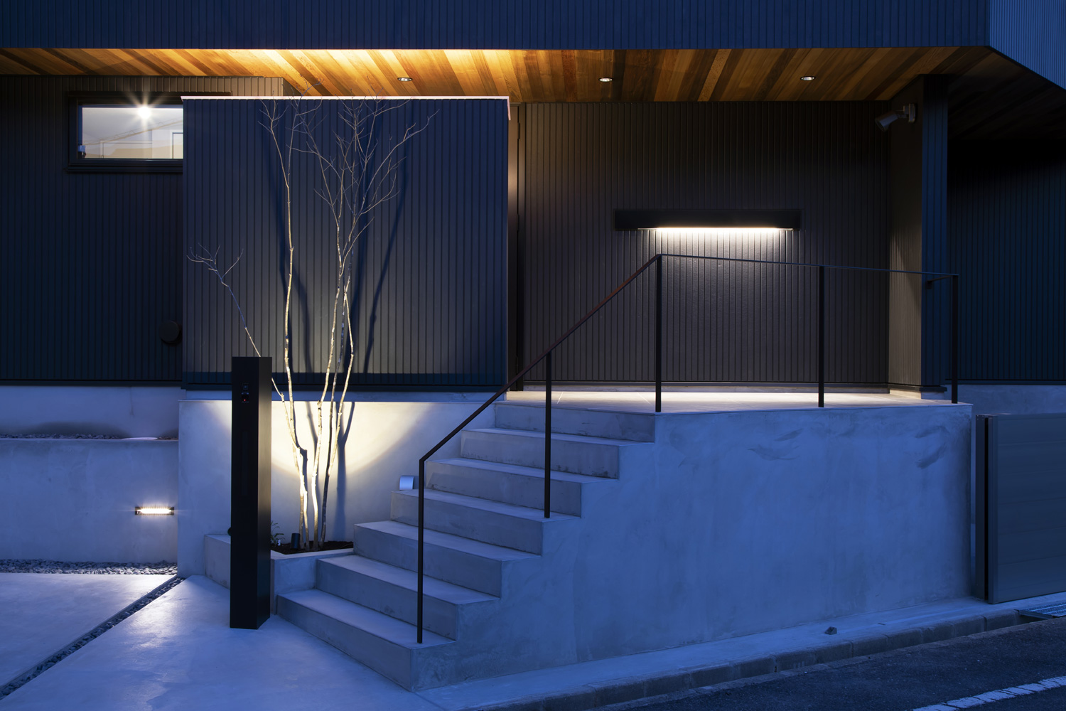 コンクリートの階段アプローチのそばにある木や玄関ポーチが照明に照らされた住宅の外観・デザイン住宅