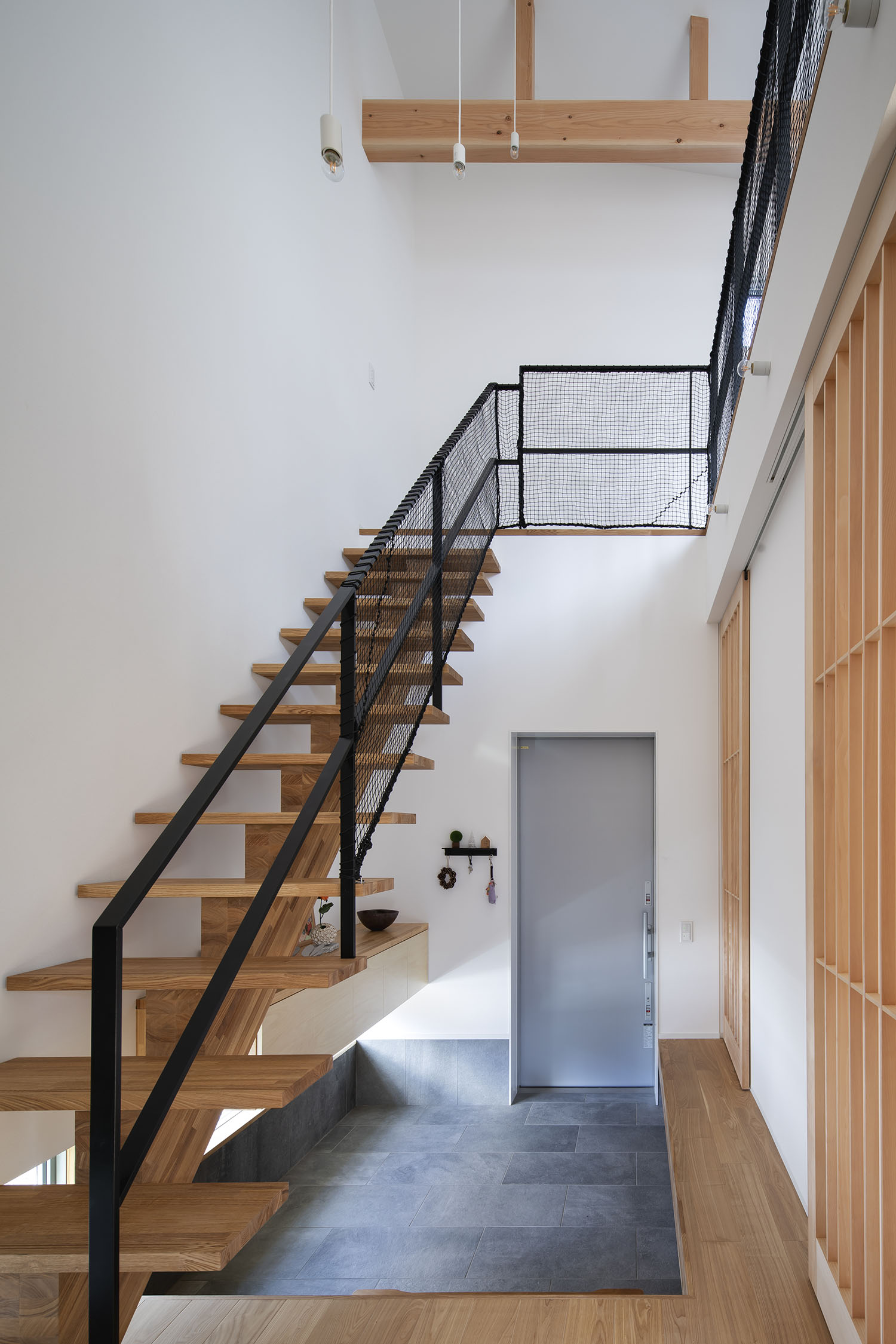 木製のスケルトン階段が横切るグレーのタイルの玄関・デザイン住宅