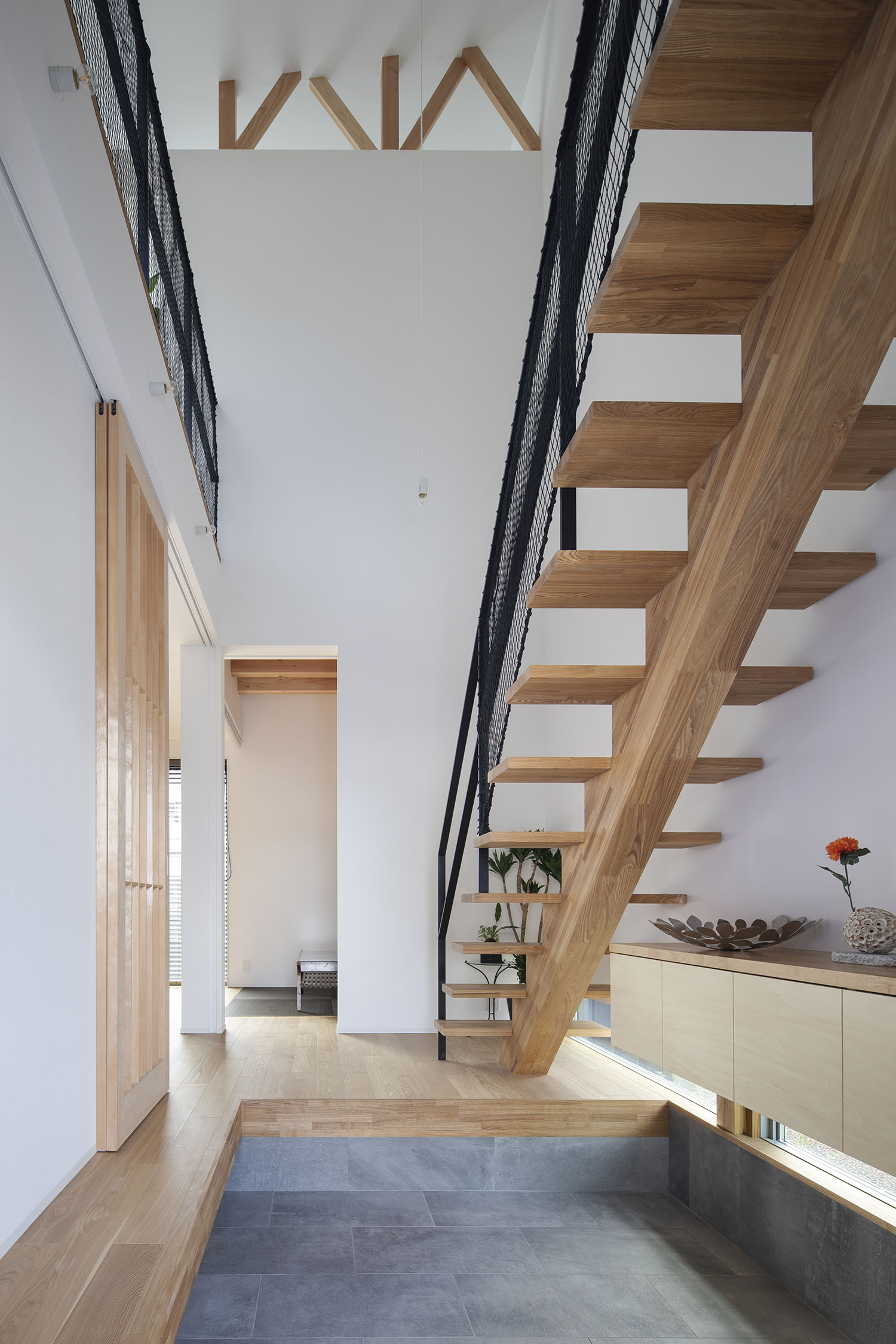 吹き抜けの玄関を横切るように木製のスケルトン階段がある様子・デザイン住宅