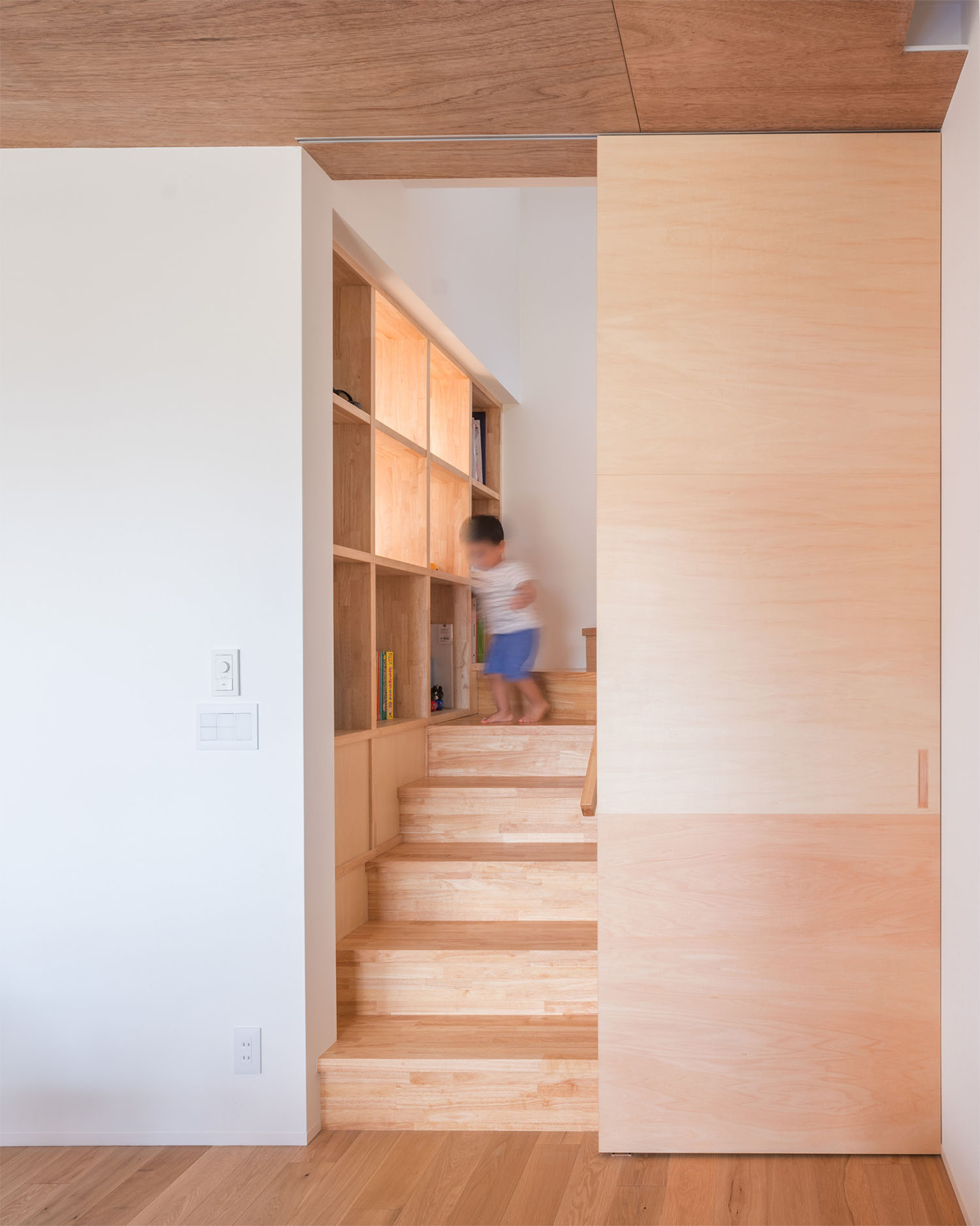 木の箱型階段のそばには壁面収納があり、子供が遊んでいる様子・デザイン住宅