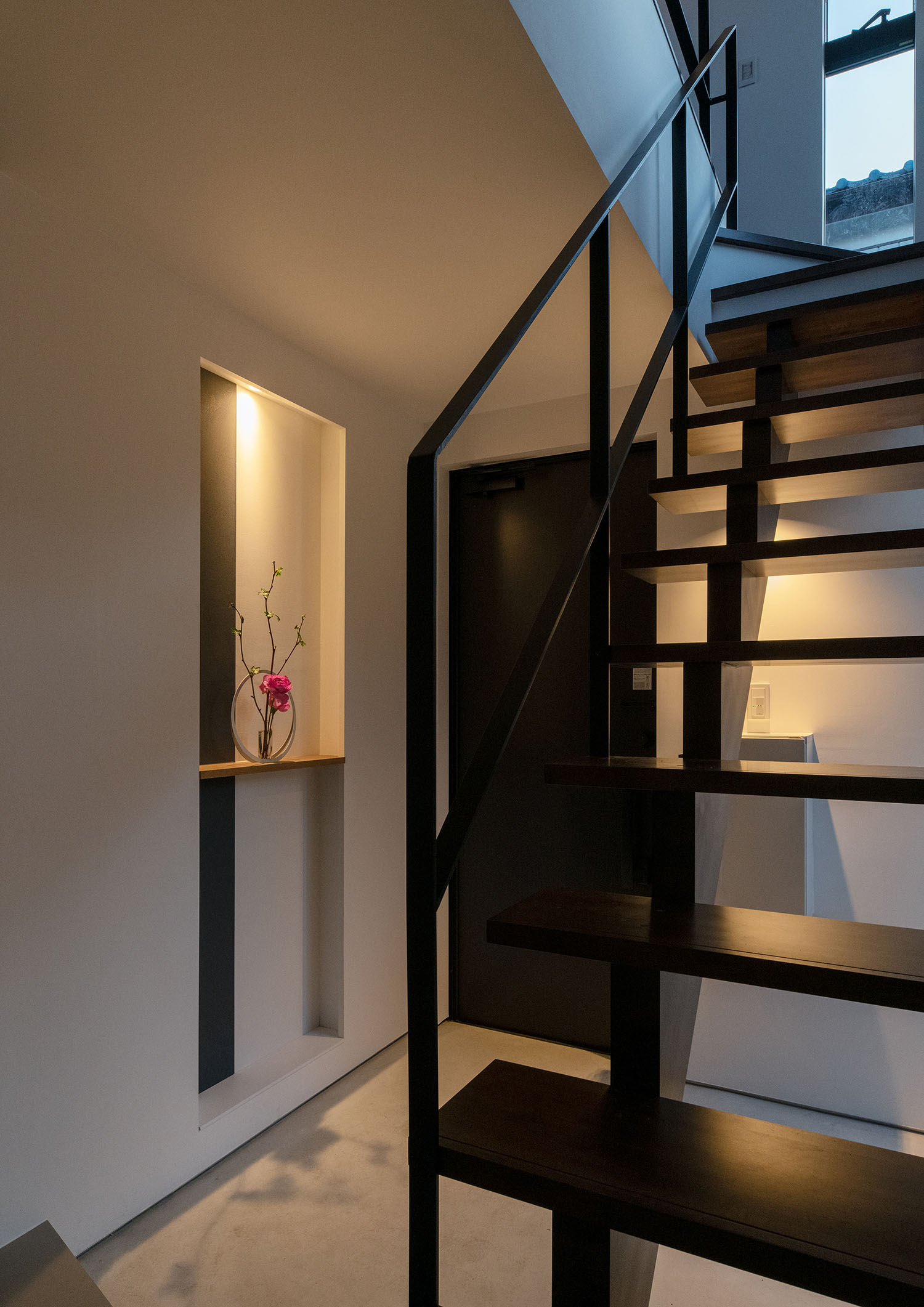 スケルトン階段越しに、飾り棚の間接照明が玄関を照らす様子・デザイン住宅