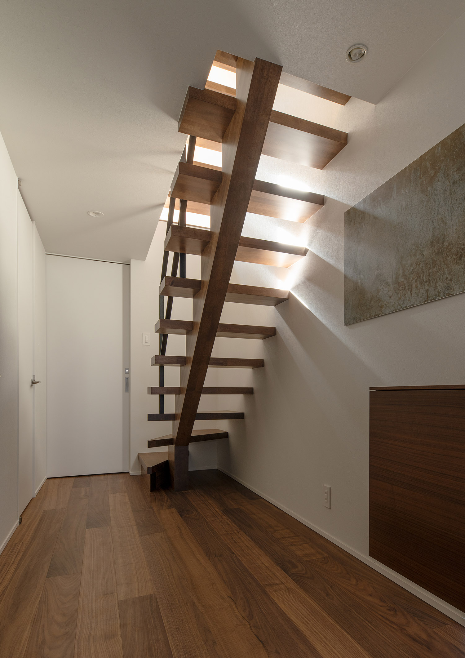 木のスケルトン階段越しに二階の光が漏れるフローリングの廊下・デザイン住宅