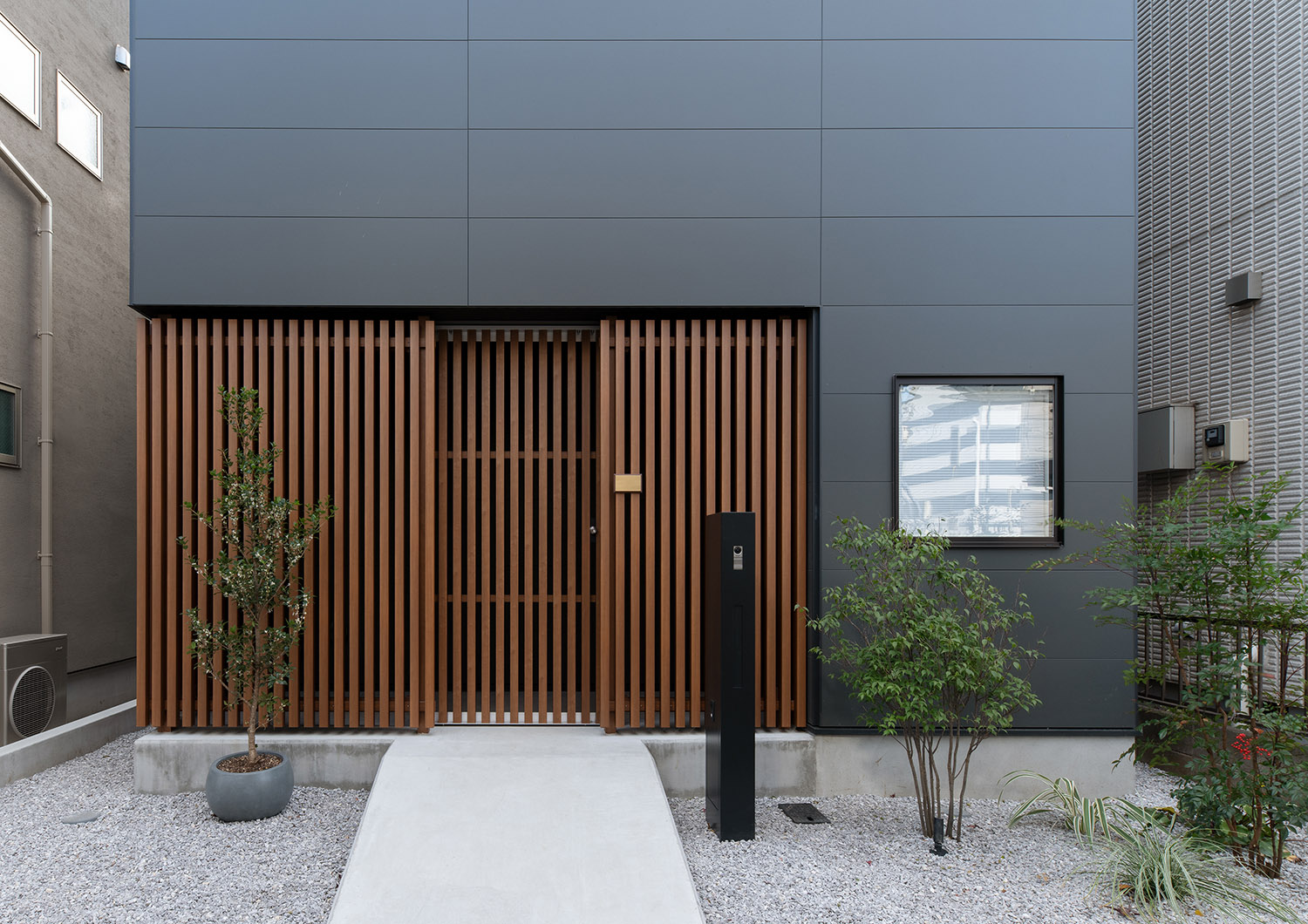 木のルーバーの壁で、砂利敷きに植栽が置かれたアプローチ・デザイン住宅