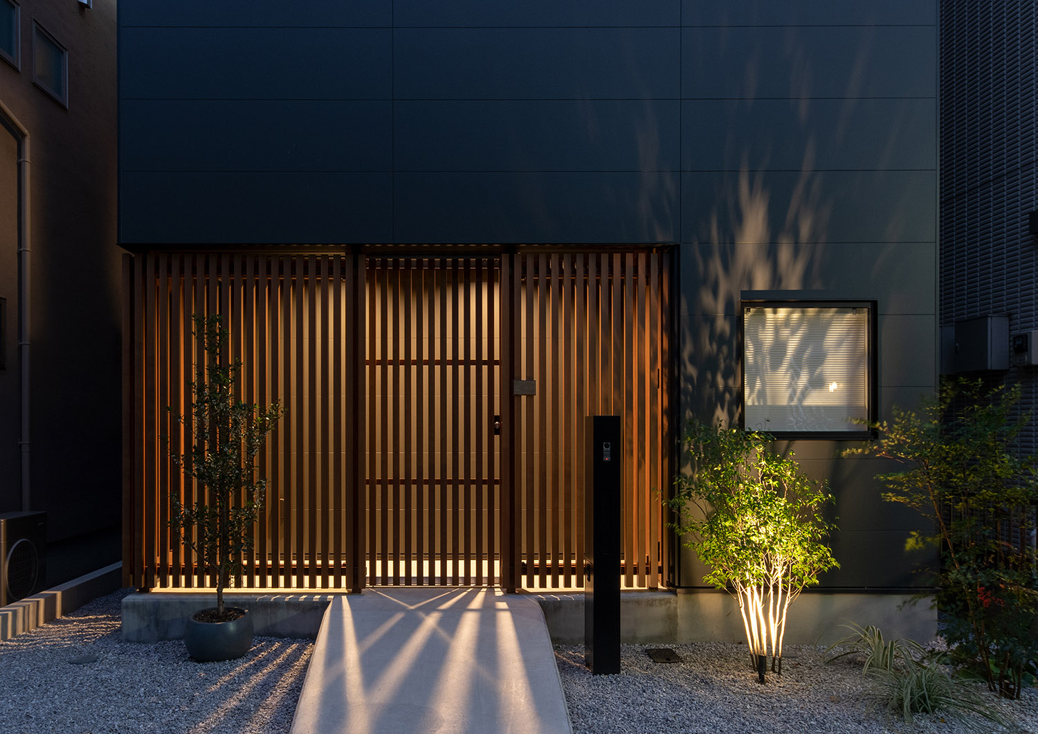 植栽が照明に照らされ、ルーバーの壁から光が漏れるアプローチ・デザイン住宅