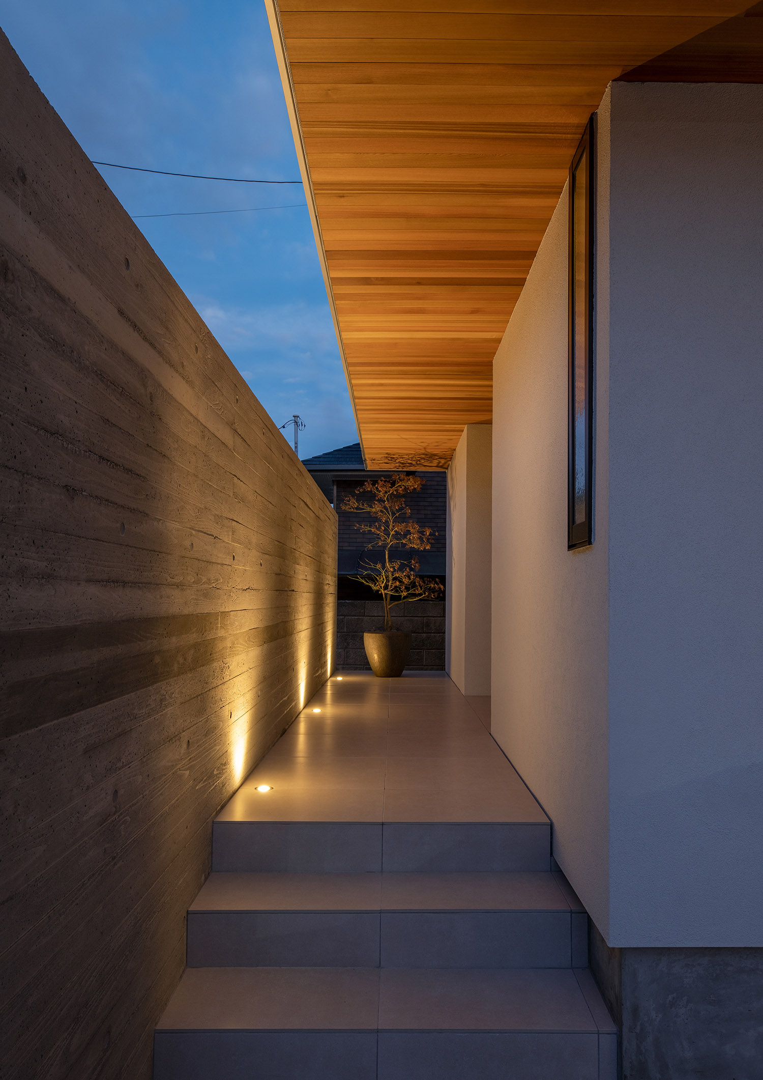 コンクリートの目隠し壁が床に埋め込まれた照明に照らされる軒天井のアプローチ・デザイン住宅