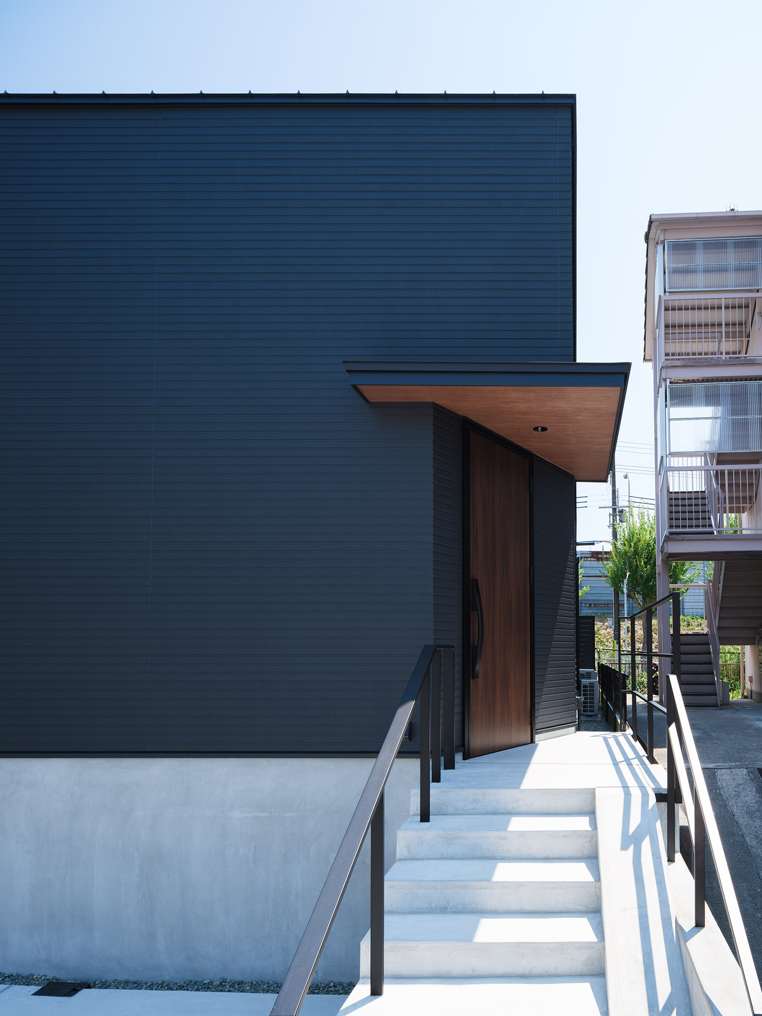 コンクリートの階段と黒い手すりがあるアプローチと、木目の庇がある玄関ポーチ・デザイン住宅