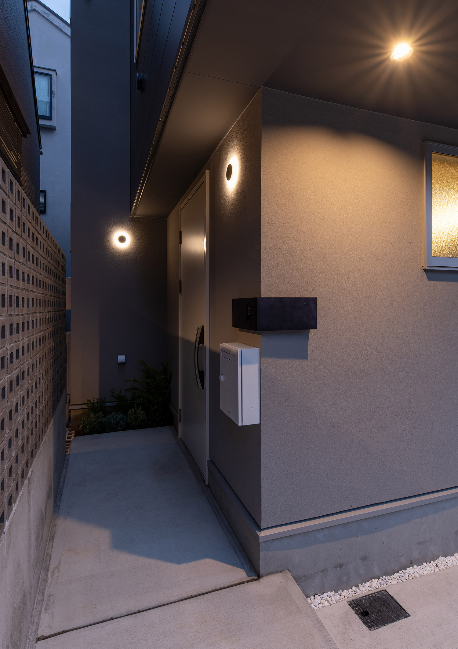 壁にシンプルな丸い照明が取り付けられたコンクリートのアプローチ・デザイン住宅