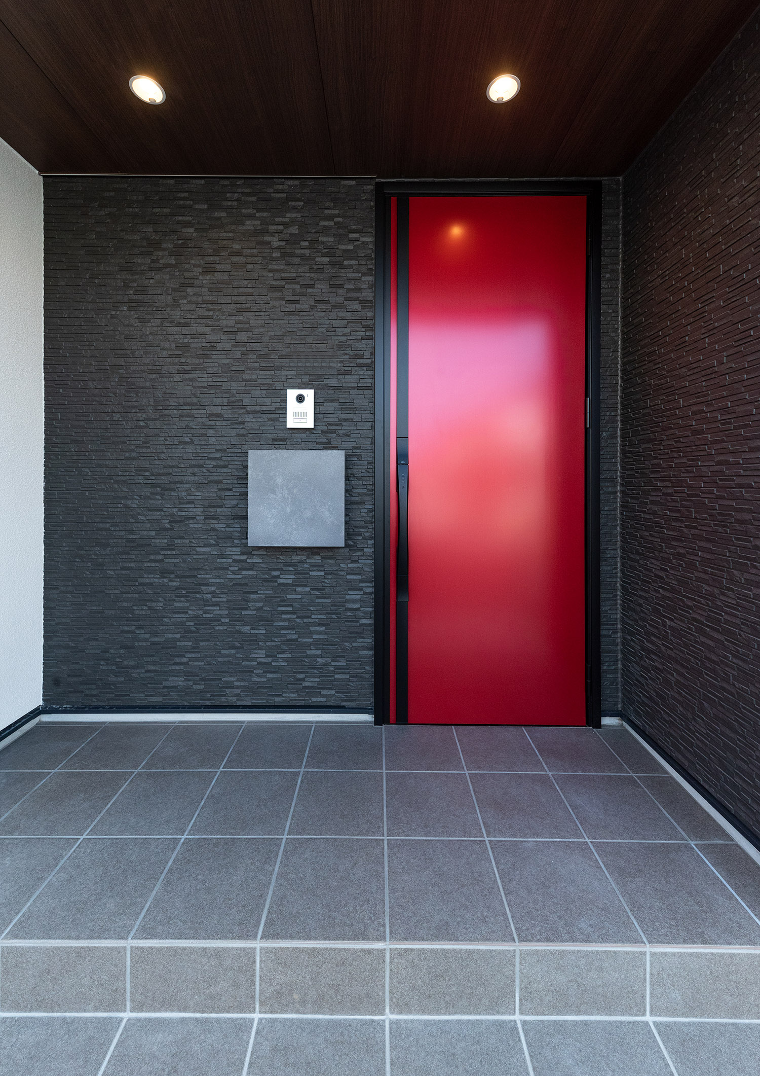 黒い壁に赤い玄関扉が取り付けられたタイル床の玄関ポーチ・デザイン住宅